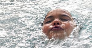 A young girl sticking her head out of the surface of pool water while swimming | Gunther Kia
