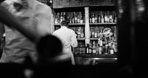 black and white image of a bar with a bartender and an assortment of alcohol bottles | Gunther Kia