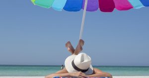A women laying on her stomach with a hat and sunglasses on at the beach underneath a colorful umbrella in Fort Lauderdale, FL | Gunther Kia